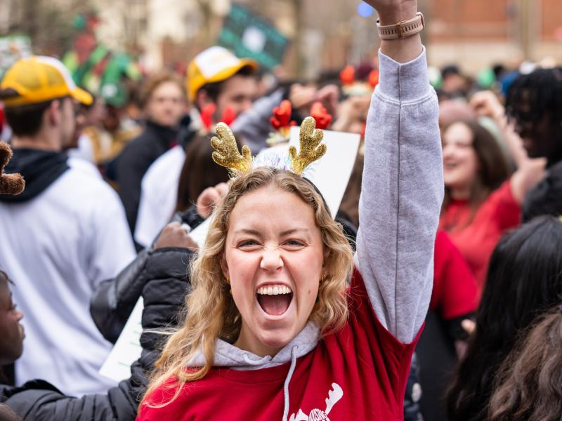 Young woman in a red hoodie and reindeer antlers cheering with her fist raised in a festive crowd outdoors.