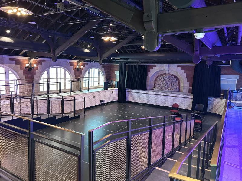 Spacious industrial-style hall with high beams, a small stage, and railings overlooking an open floor.