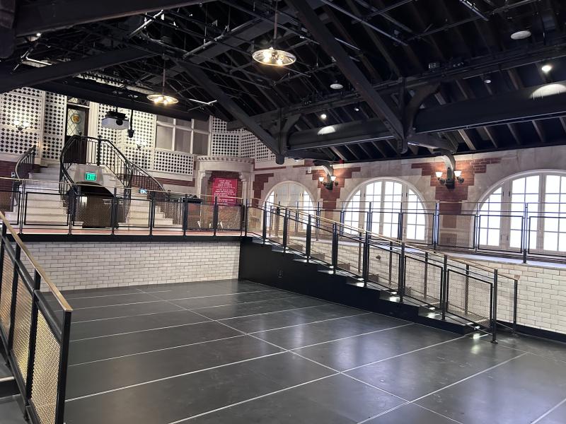 Large open hall with black flooring, arched windows, and metal railings under exposed beams.