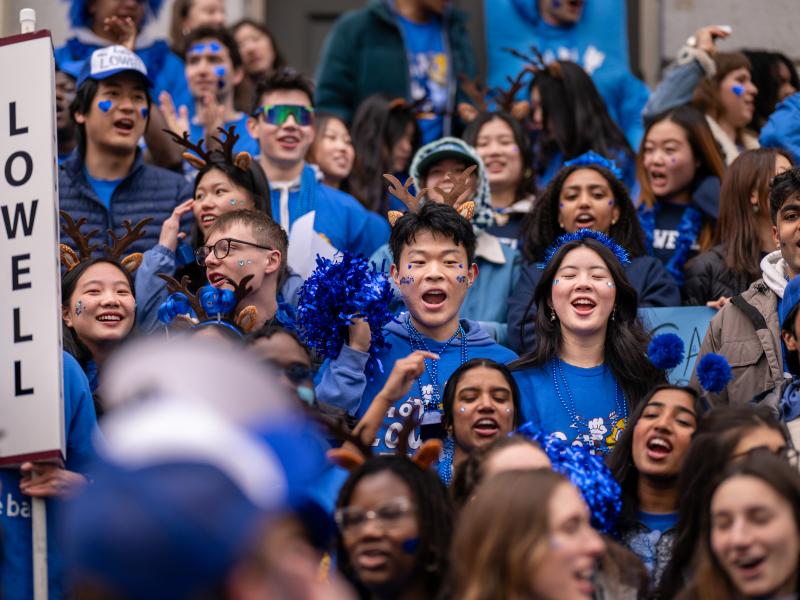 Crowd of students in blue clothing and face paint cheer, holding a vertical “LOWELL” sign.