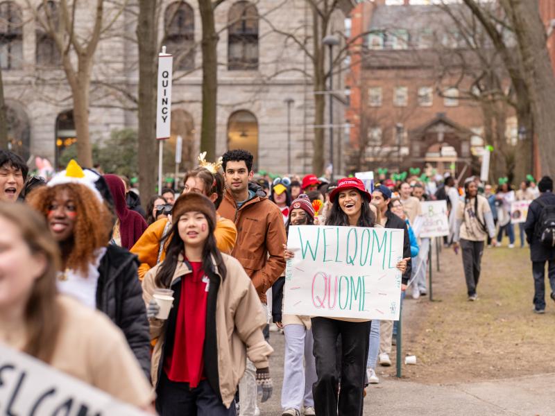 Students march through campus holding signs, including one that reads “WELCOME QUOME,” during a festive outdoor event.