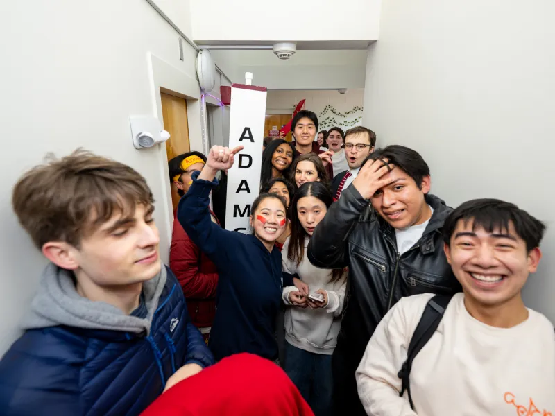 Excited students crowd a hallway, smiling and cheering around an “Adams” House sign.