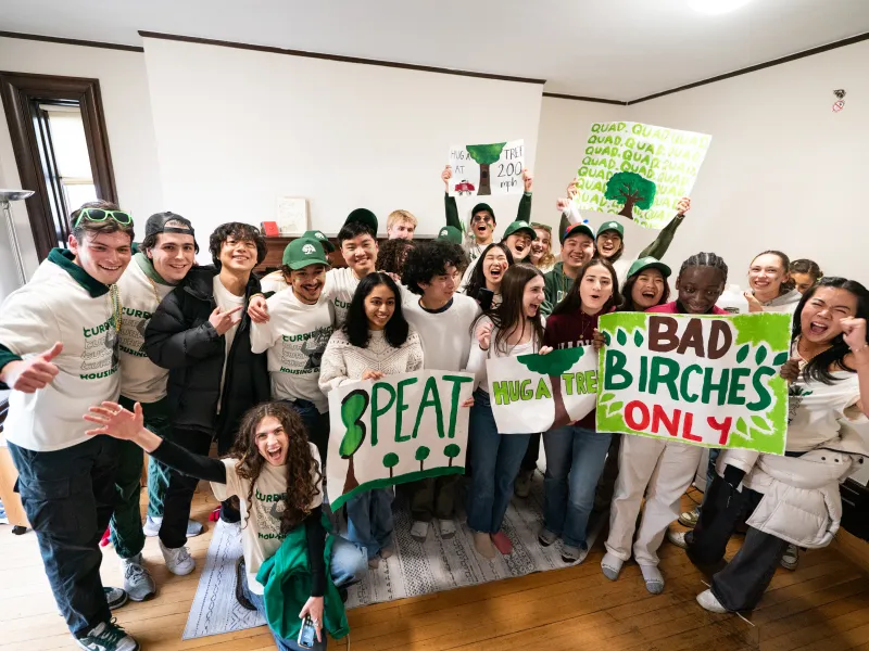 Group of students in green and white shirts cheer indoors, holding handmade tree-themed signs.