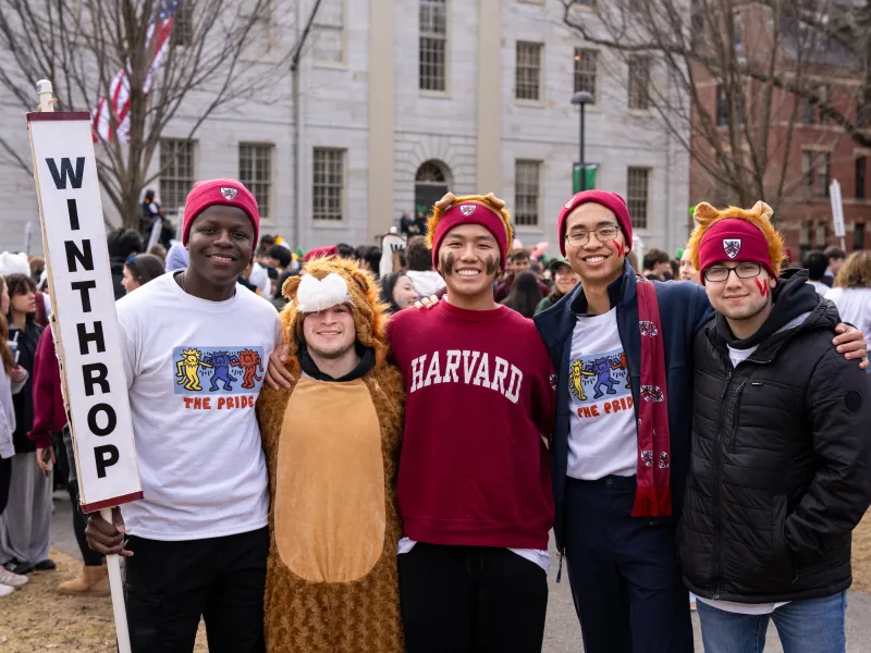 Five students in Harvard gear pose together outdoors, one holding a “Winthrop” house sign.