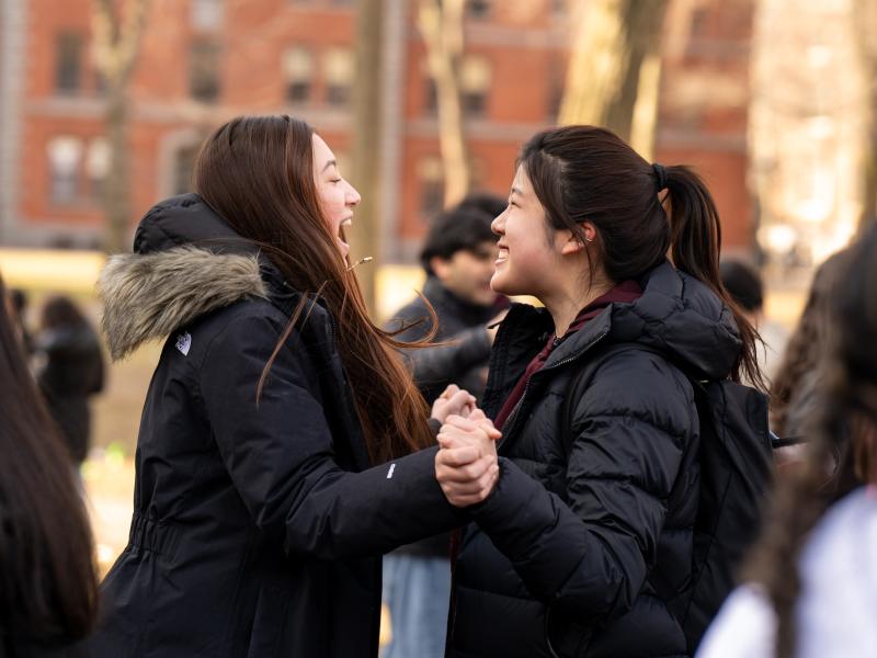Two students in winter coats laugh and hold hands while celebrating outside on campus.
