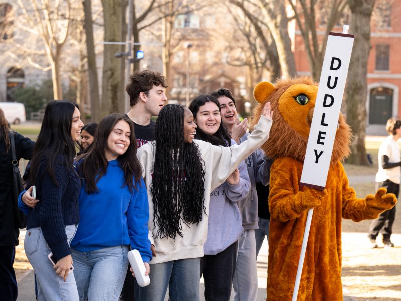 Students laugh and high-five a lion mascot holding a “Dudley” sign in a campus yard.