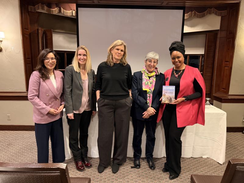 Five women stand together smiling in front of a projector screen, one holding the book “Outspoken.”