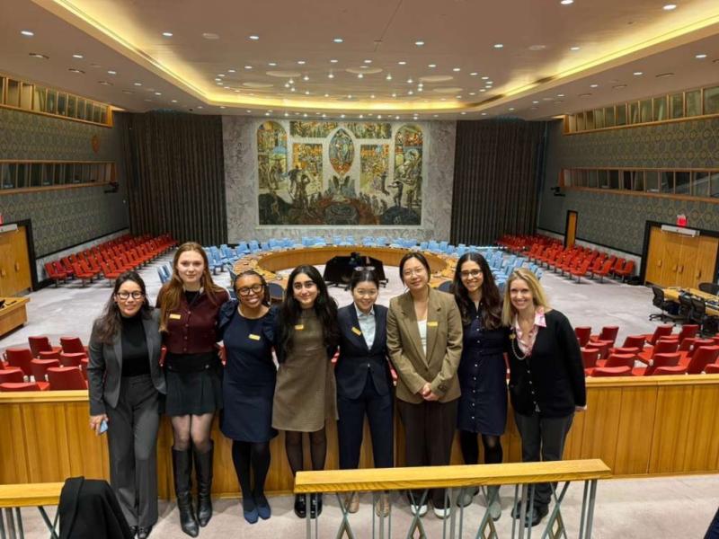 Group of students pose together in the UN Security Council chamber with empty seats behind them.