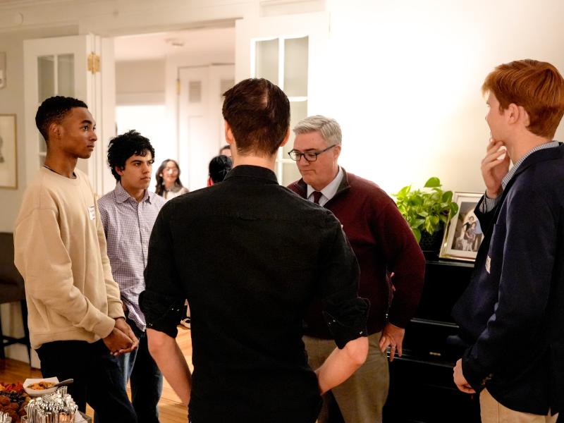 Small group of students and an older adult talk together at a casual indoor reception.