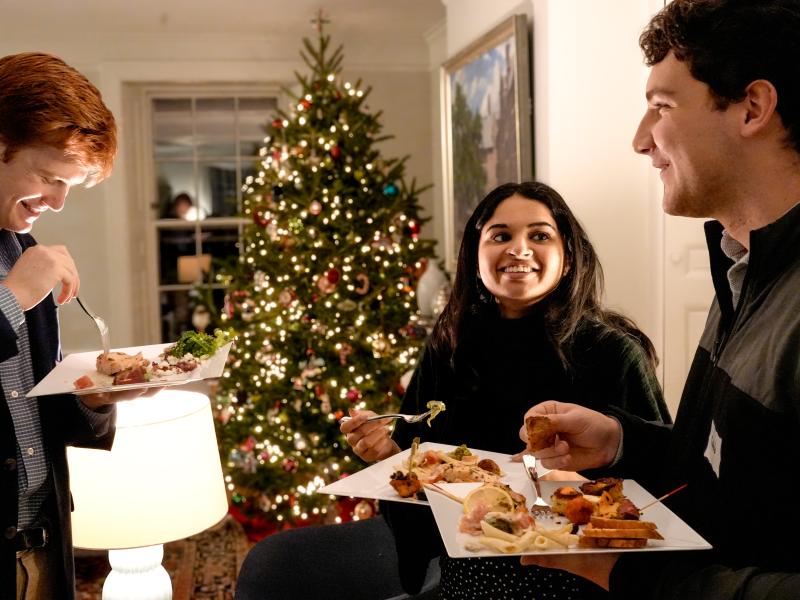 Three students laugh and eat holiday food in front of a decorated Christmas tree.