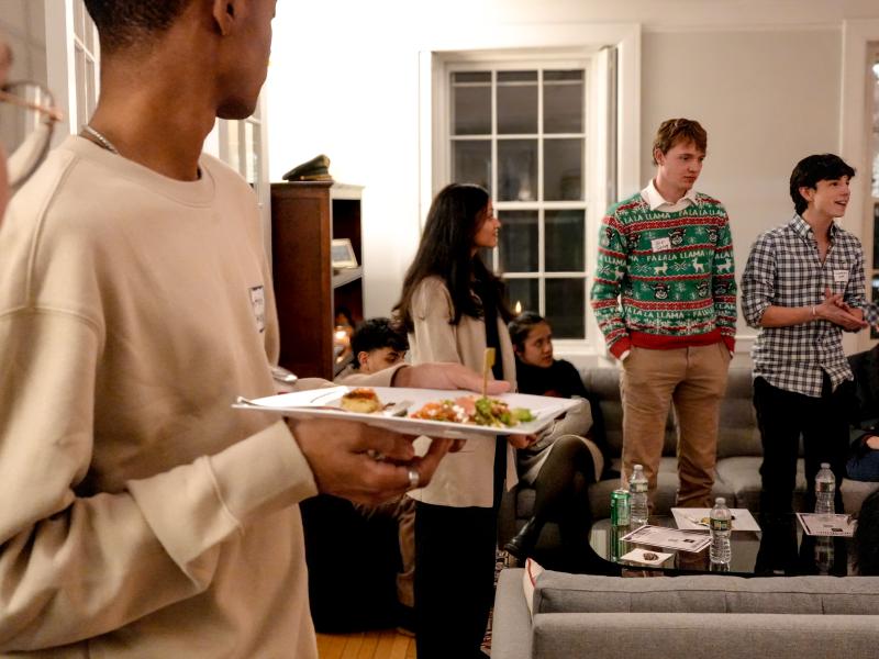 Students gather in a cozy living room, some standing and speaking while others sit and listen during a holiday event.