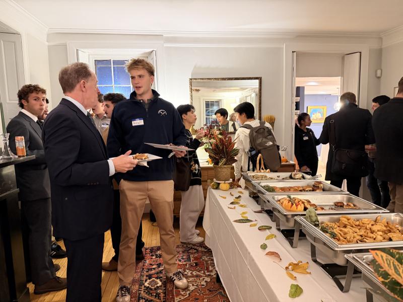 Guests in semi-formal attire mingle and eat beside a long buffet table at an indoor reception.