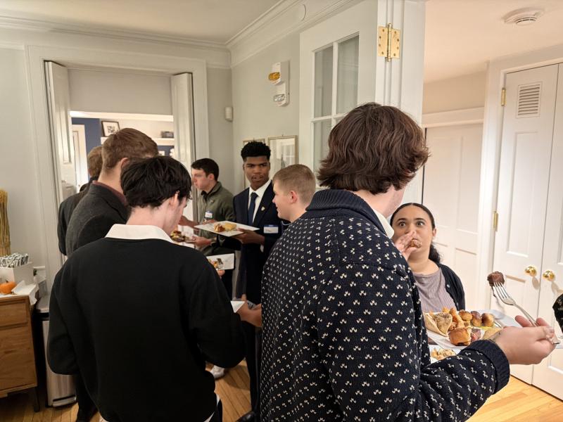 Students gather in a hallway holding plates of food and chatting at a reception.