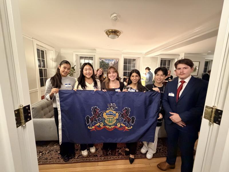 Group of students stand in a living room holding a Pennsylvania state flag.