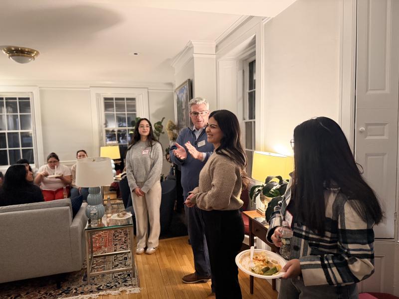 Students and a faculty member gather in a living room, one person speaking while others listen and hold plates of food.