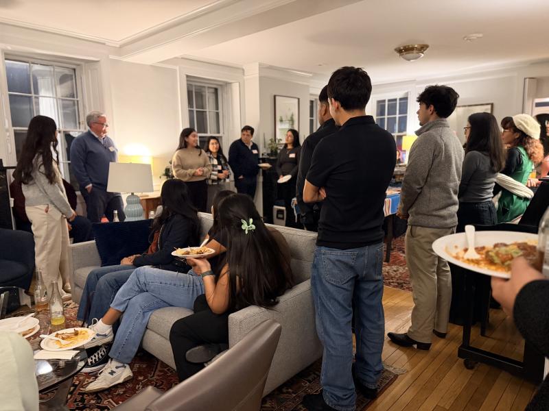 Students gather in a living room, standing and sitting with plates of food while one person speaks.