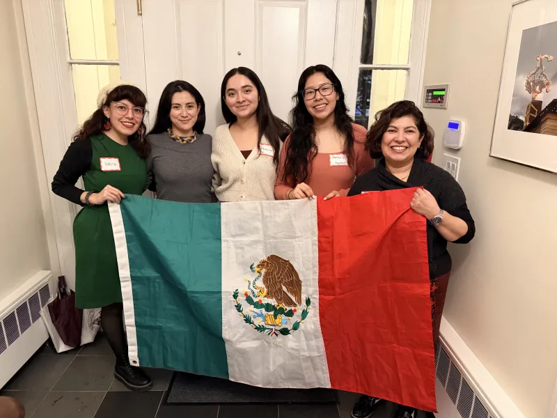 Five women smile while holding a large Mexican flag in a hallway.