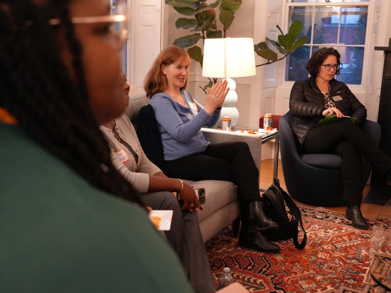 Students and staff sit in a cozy living room, one woman speaking while others listen and snack.