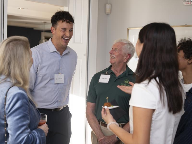 Students and alumni stand in a small circle at a reception, laughing and talking.