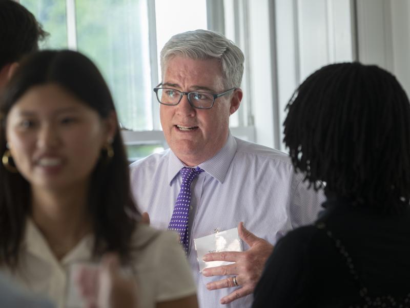 A man with grey hair, glasses, and a purple tie speaks animatedly while gesturing during a conversation with students at an indoor event.