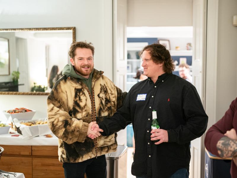 Two men shake hands in a room with a buffet table; one wears a fleece jacket with a wolf print, and the other wears a black button-down shirt and a name tag.