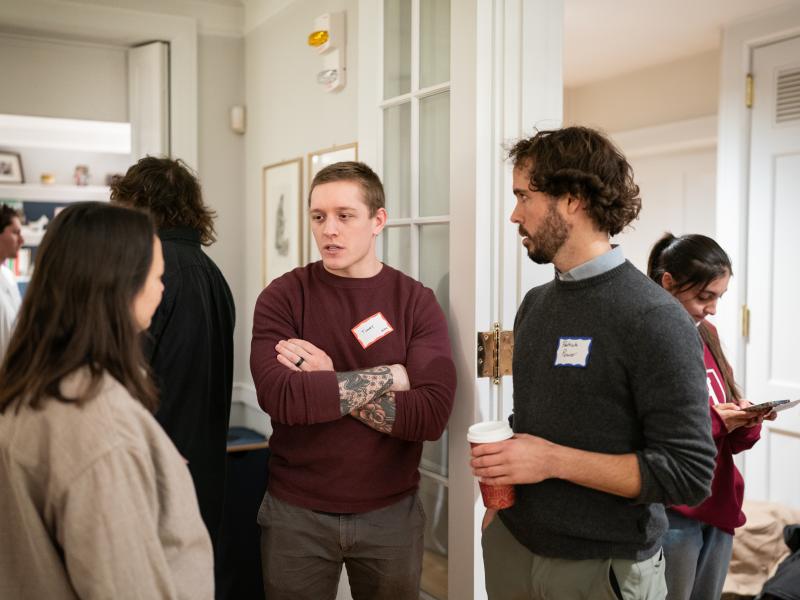 A man with arm tattoos and a maroon sweater speaks with a group of people at an indoor event, while another man in a grey sweater holds a coffee cup and listens.