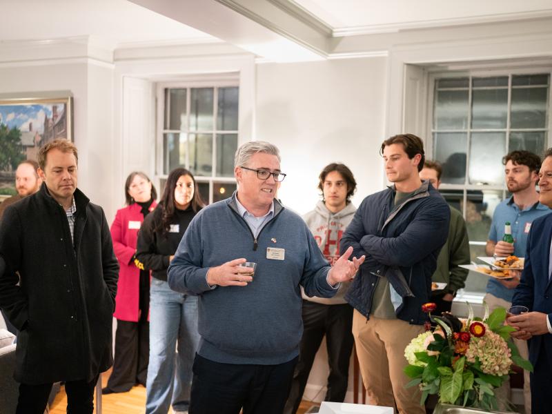 A man in a blue quarter-zip sweater speaks and gestures to a group of attentive guests during an indoor gathering.