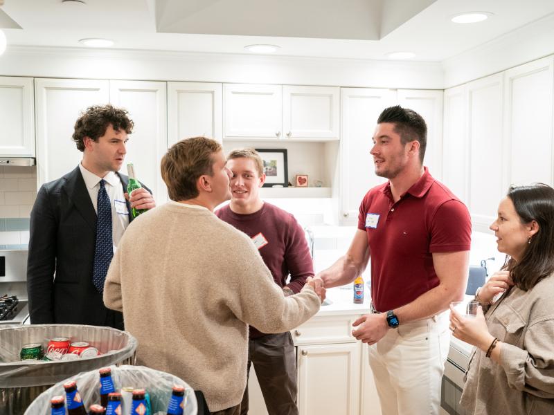 A group of people chat in a kitchen; two men shake hands while others look on holding drinks.