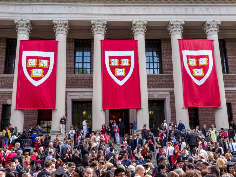 Large red banners with the Harvard University shield hang between stone columns on the front of a brick campus building.