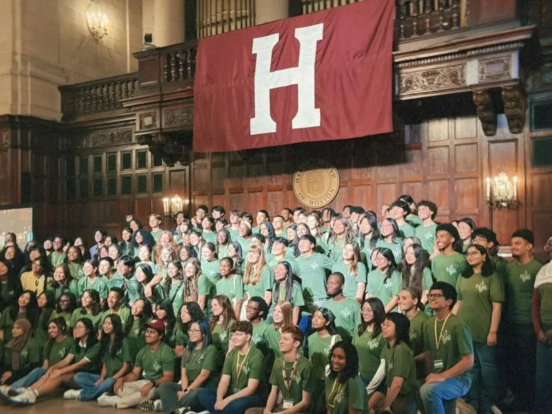 A large group of diverse students wearing matching green t-shirts posing for a group photo inside a grand, wood-paneled hall. They are gathered beneath a large crimson banner displaying a white letter "H" and a circular Harvard Club of Boston emblem.