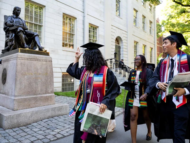 A female student in regalia tips her hat to the John Harvard statue as she walks by.