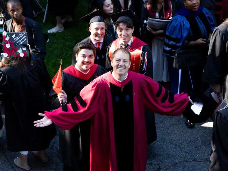 Danoff Dean of Harvard College, David Deming, with three male students behind him, smiling upward at the camera while wearing their regalia.