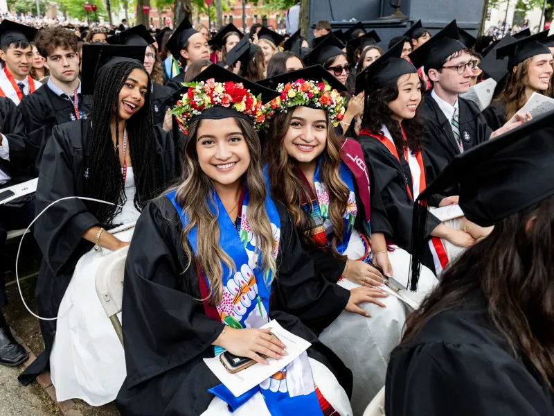 Two female graduates seated next to each other and smiling while wearing colorful regalia and flower crowns on their caps.