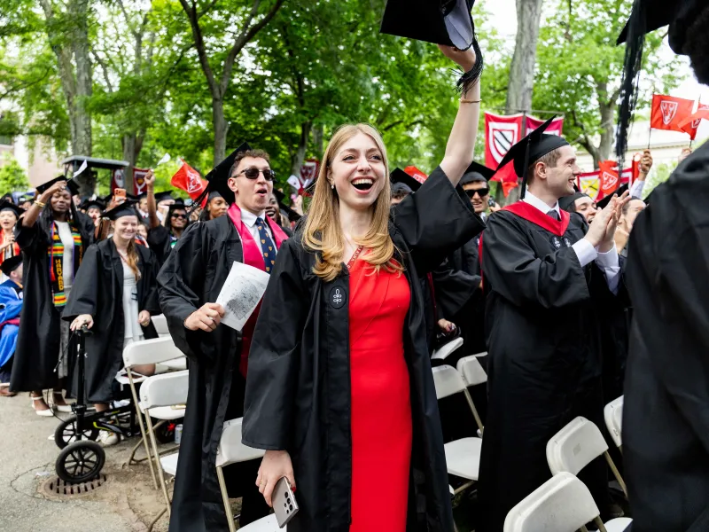 A young female graduate in a red dress holding her cap high above her head.