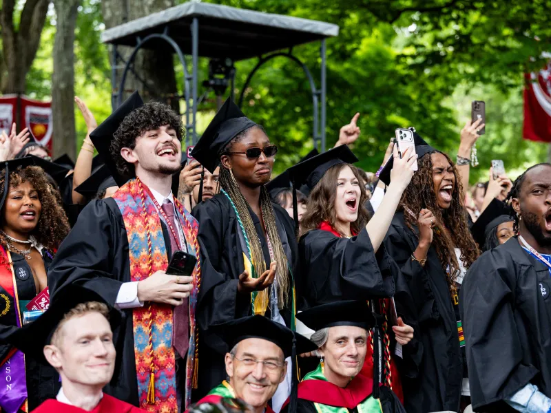 A group of 6 graduates standing up from their seats and cheering during the ceremony.