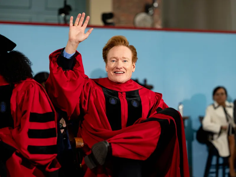 Conan O'Brien waving to the crowd while seated onstage during the Harvard Commencement.