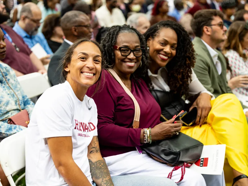 Harvard Women's Basketball Coach, Carrie Moore, seated and smiling at one of the House ceremonies next to two other women.