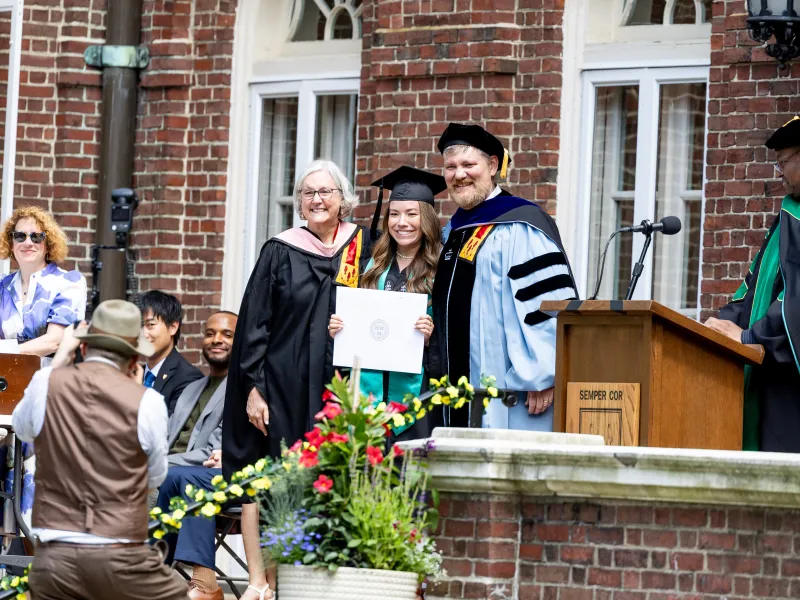A female student holding up her degree and smiling for a picture with two faculty members beside her.