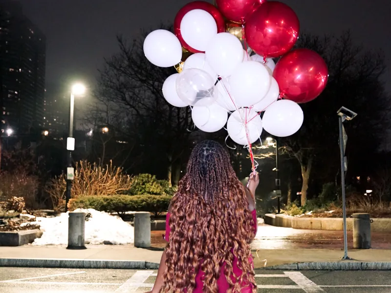 Photo of girl in red dress holding red and white balloons while walking across the street