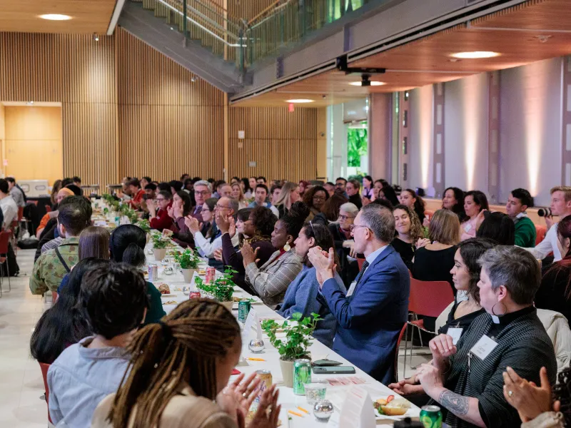 Many students, faculty, and staff sit at several long tables, eating and talking