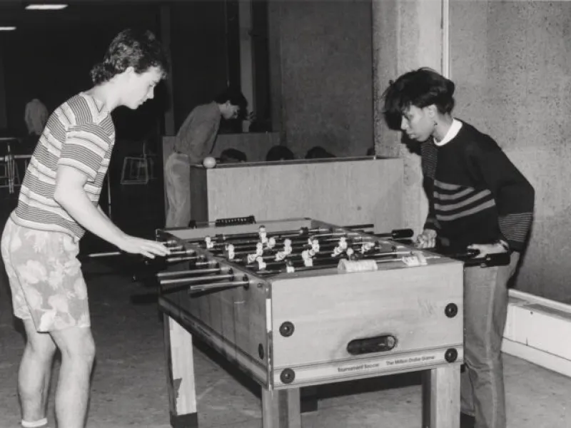 A black and white image of two male students playing on a fooseball table inside Currier House in 1987.