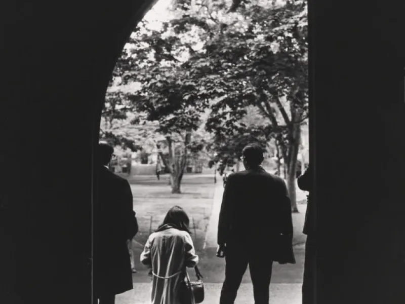A black and white photo from 1960 of students passing under the arch into Harvard Yard.