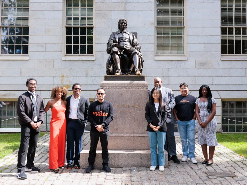 A group of 8 visitors posing with the John Harvard statue outside of University Hall.