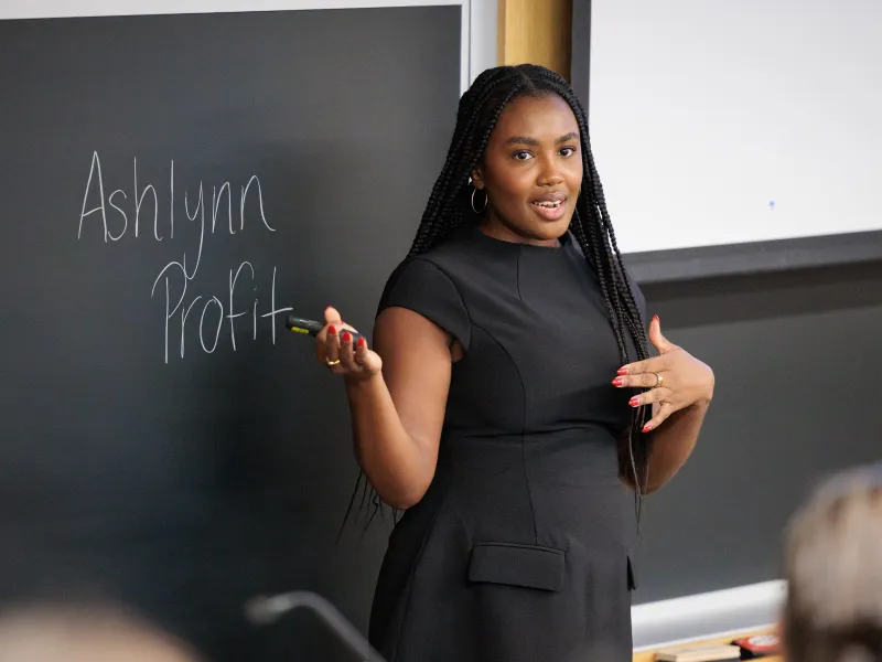 Ashlynn Profit speaking while standing in front of a blackboard with her name written in white chalk on it.