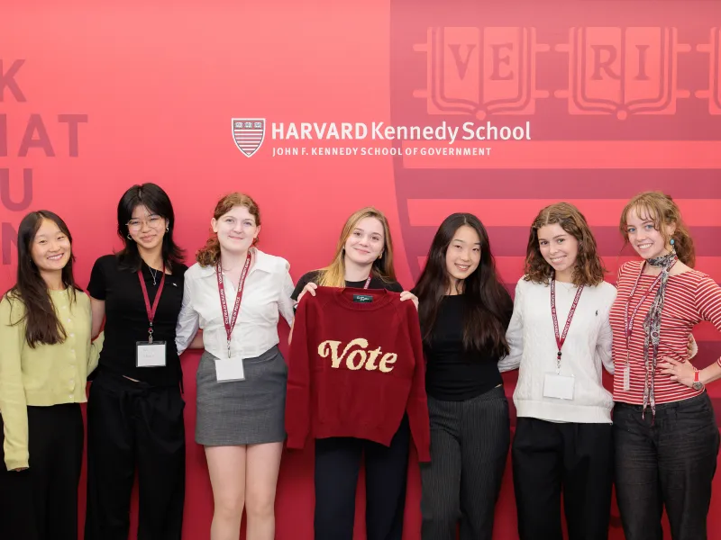 A group of 7 female students smiling in front of a red wall that reads "Harvard Kennedy School John F. Kennedy School of Government."