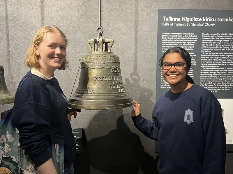 Students Živa Benedejčič '28 and Mirika Jambudi '27 smiling next to a bell of St. Nicholas Church.