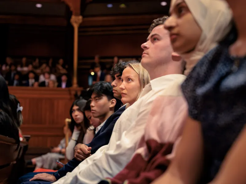 A group of students sitting in a line and looking up to the stage intently.