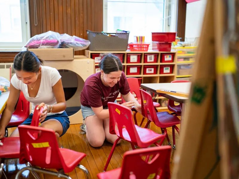 Two female students bent down working on chairs in a classroom.