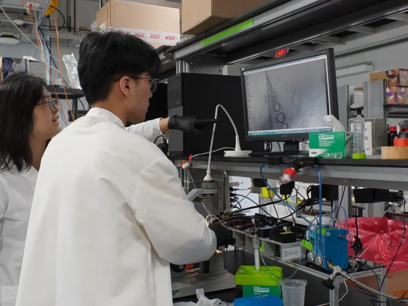 Two people in lab coats in 2026 looking at a computer in their lab.
