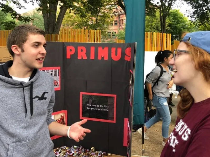 two people talking outside next to a poster that says Primus